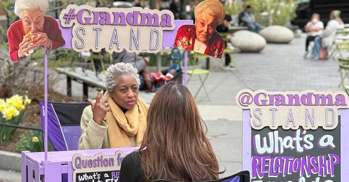 A grandma sits at a purple lemonade stand talking to a stranger. It's called "The Grandma Stand"