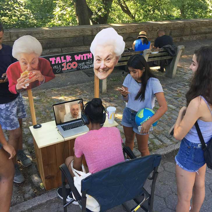 A group of teenagers sits at a pop-up stand in a park in New York, speaking to a grandma on a laptop