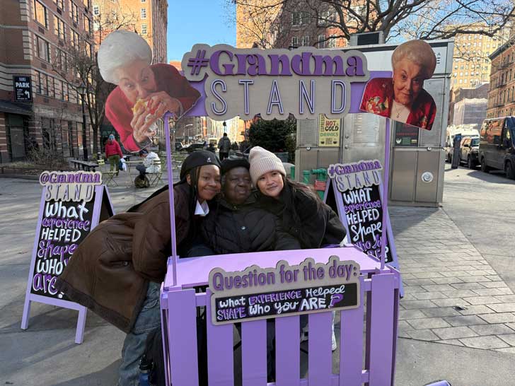 Two teenagers pose for a photo with an old woman at a Grandma Stand in New York City
