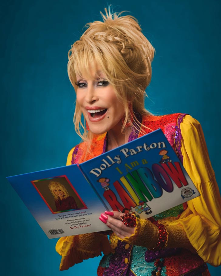 Dolly Parton wears a colorful jacket, smiling while holding a children's book: "I Am A Rainbow"