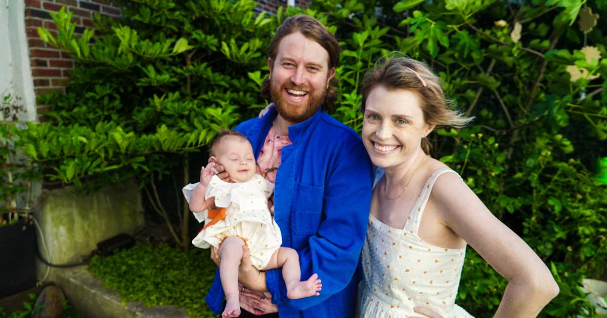 A husband, wife, and baby stand in front of a green, leafy wall, smiling. They are Andrew Rose Gregory and Casey McIntyre.