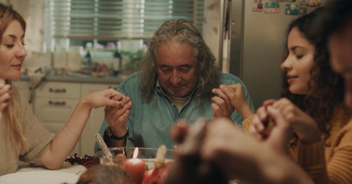 Family seated around a dinner table holding hands and praying, with an older man at the center, a lit candle and holiday food in the foreground, and a cozy kitchen in the background.