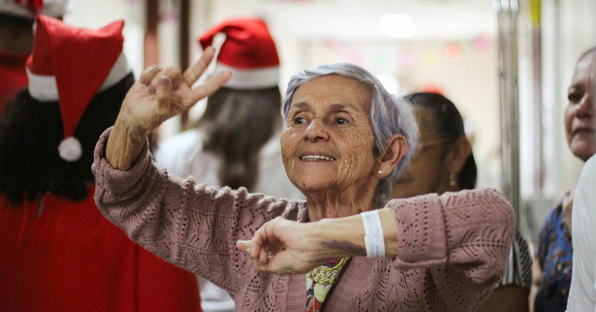 An elderly woman dances in the hallway of a nursing home, surrounded by people in Santa hats