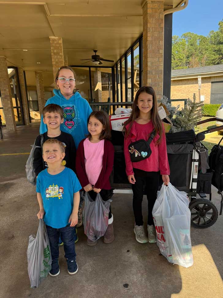 Five children stand outside of a nursing home with small Christmas trees and bags of holiday decorations