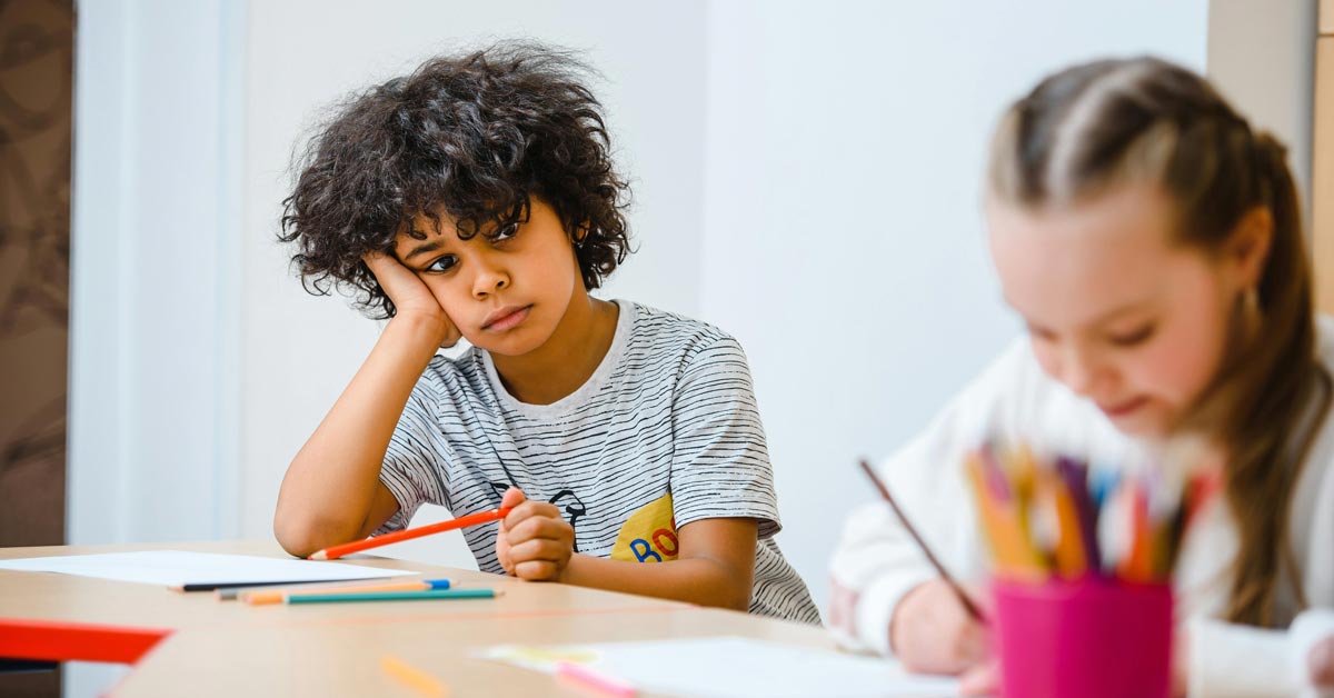 A child sits at a desk, bored, resting his head on his hand