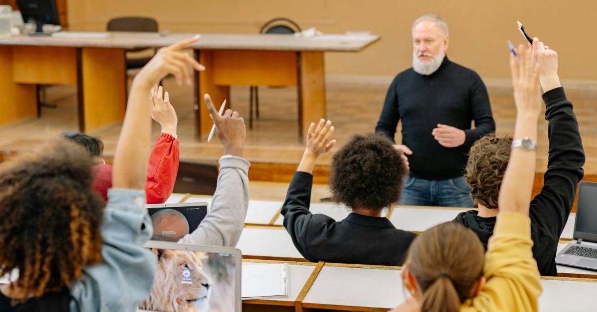 A group of students raising hands in class as a teacher leads. 