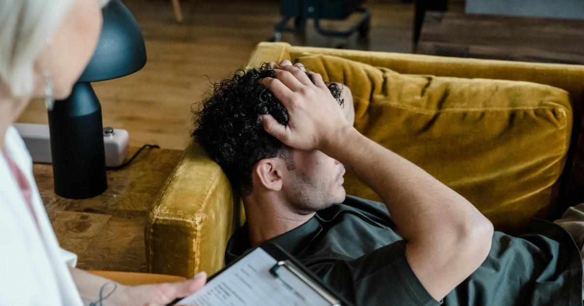 A man laying down on a couch holds his head as a therapist sits just out of frame