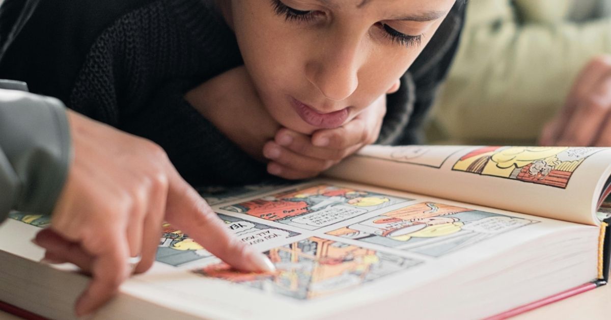 A young boy reads a comic book as a hand points along a page.