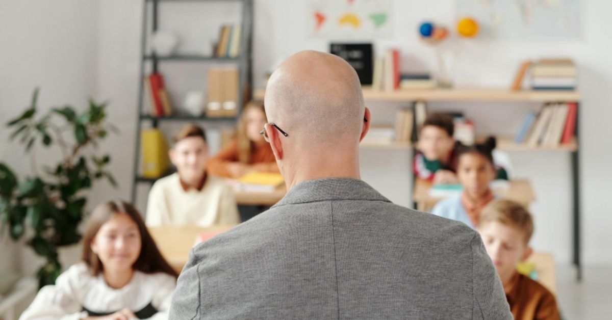 A bald-headed teacher stands at the front of the classroom (his back facing the camera) as young children smile up towards him