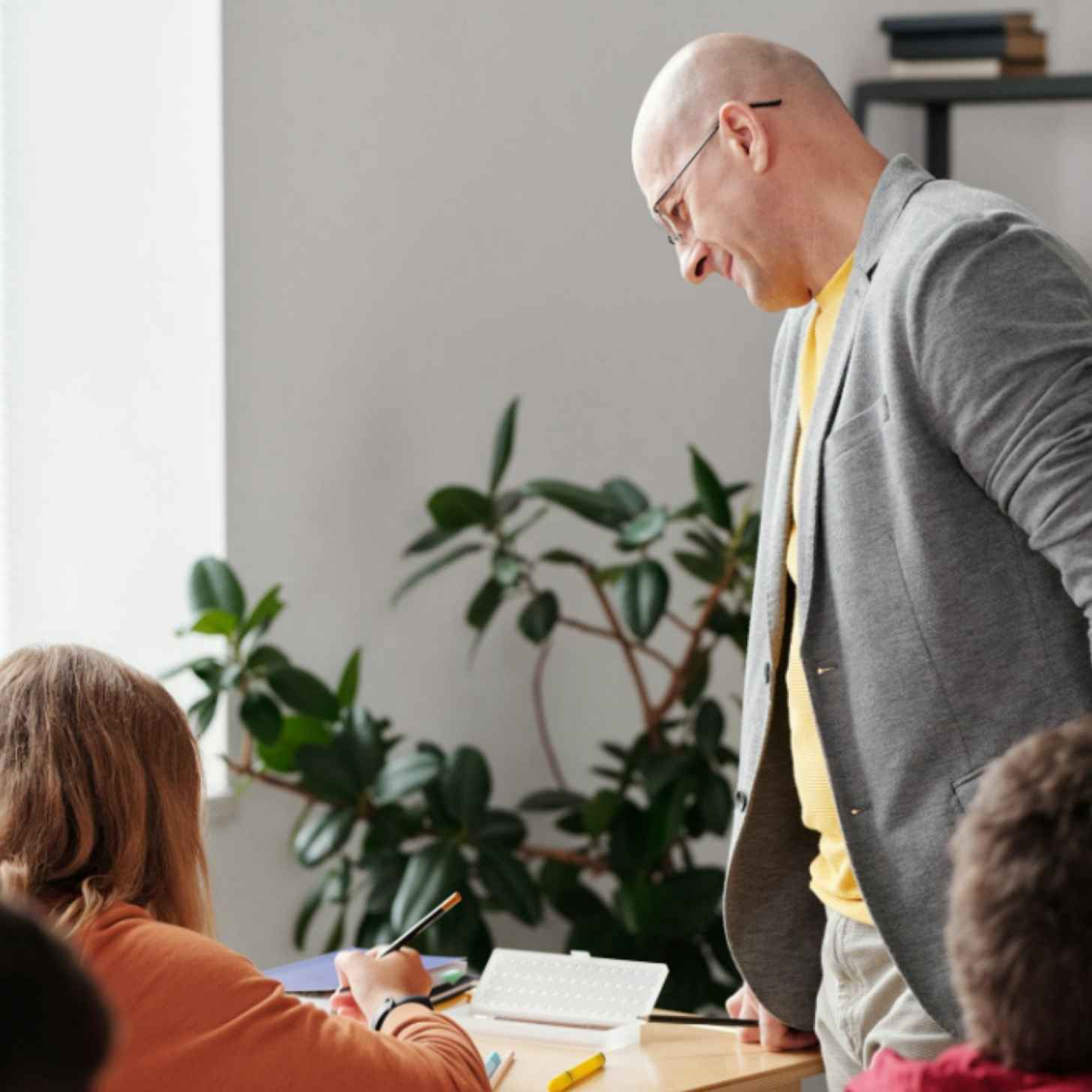 A bald-headed man (a teacher) smiles down at his students as he walks around a classroom checking their work