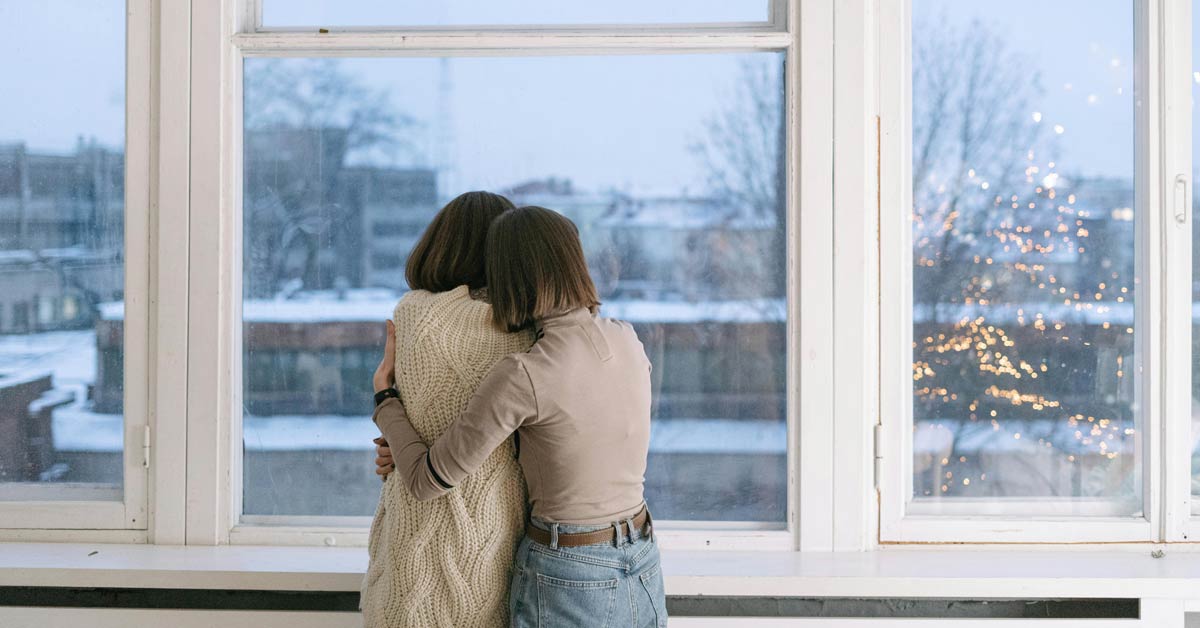 Two women, seen from the back, hug in front of a window. Outside, snow is falling.