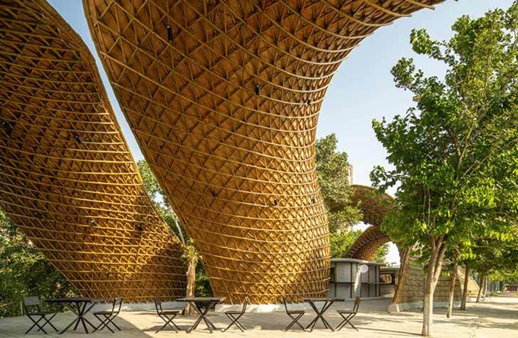 The Hungry Caterpillar -- a bamboo structure winding around kitchen stalls printed from concrete. This is a wide shot that shows how tall the bamboo reaches. Three tables and 6 chairs are in the foreground. 