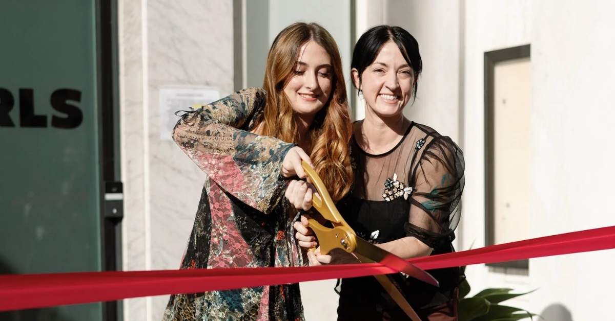 A teenage girl and an adult woman smile as they cut a red ribbon with oversized gold scissors outside a community space.