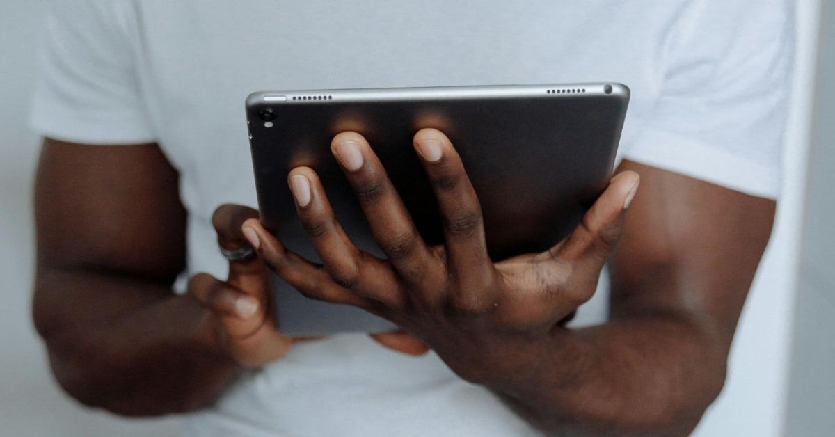 A muscular Black man wearing a white t-shirt holds a tablet in both hands 