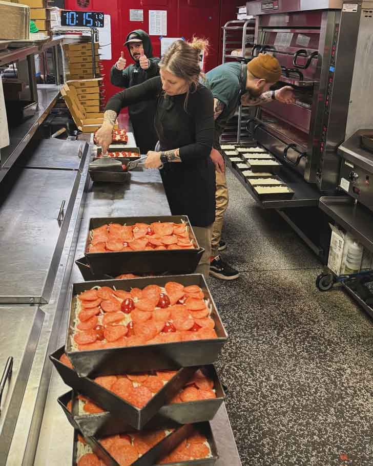 Three people in a commercial kitchen make pizzas