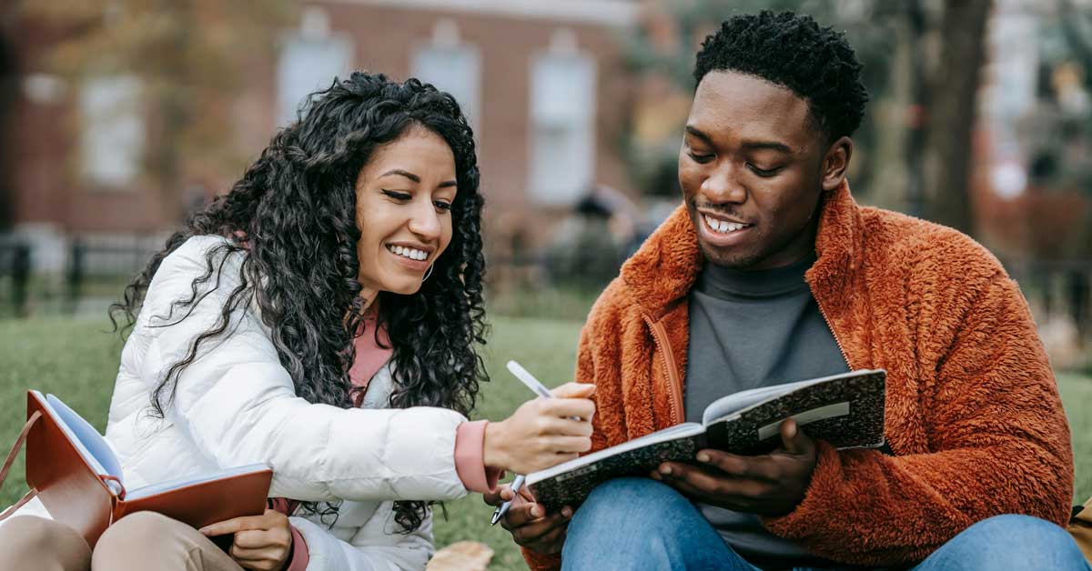 Two college-aged people, a woman and a man, sit in a courtyard, smiling into a notebook