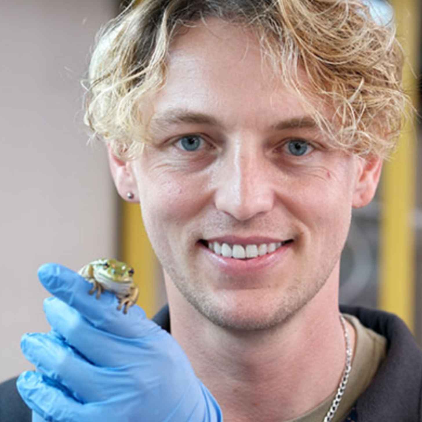 Anthony Waddle, a young man with blond hair, holds a green and golden bell frog in his gloved hands in a lab as he smiles