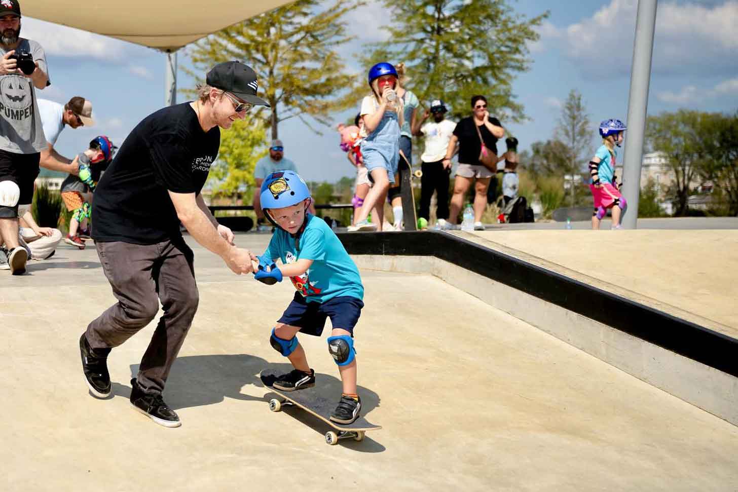 An adult helps a child learn how to skateboard at a skatepark, surrounded by other children