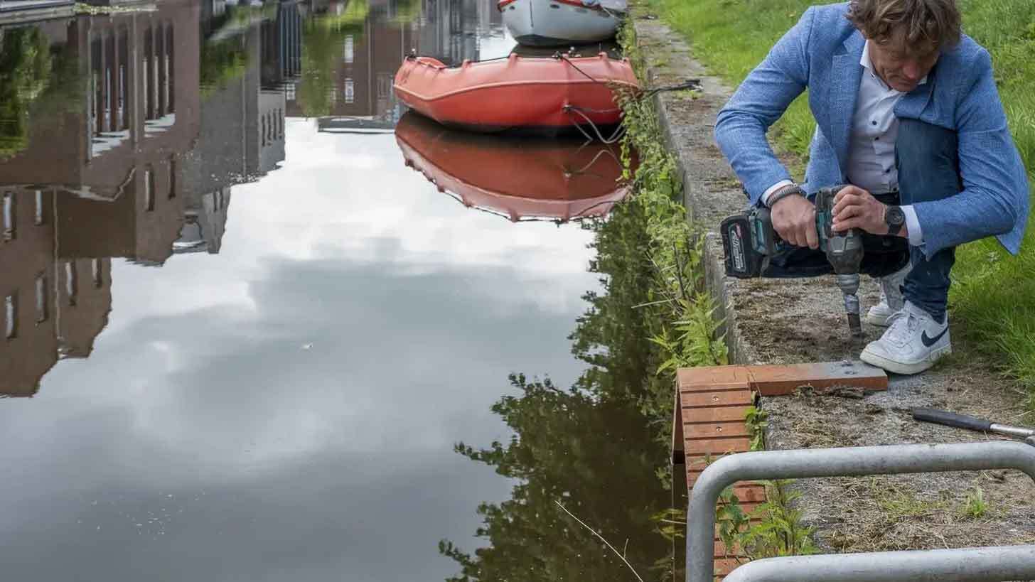 A middle-aged blonde man wearing a blue suit jacket and jeans drills into a tiny wooden staircase along a waterway.