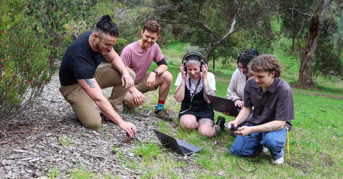 A group of researchers kneel outdoors over a patch of soil, listening with headphones and holding a laptop