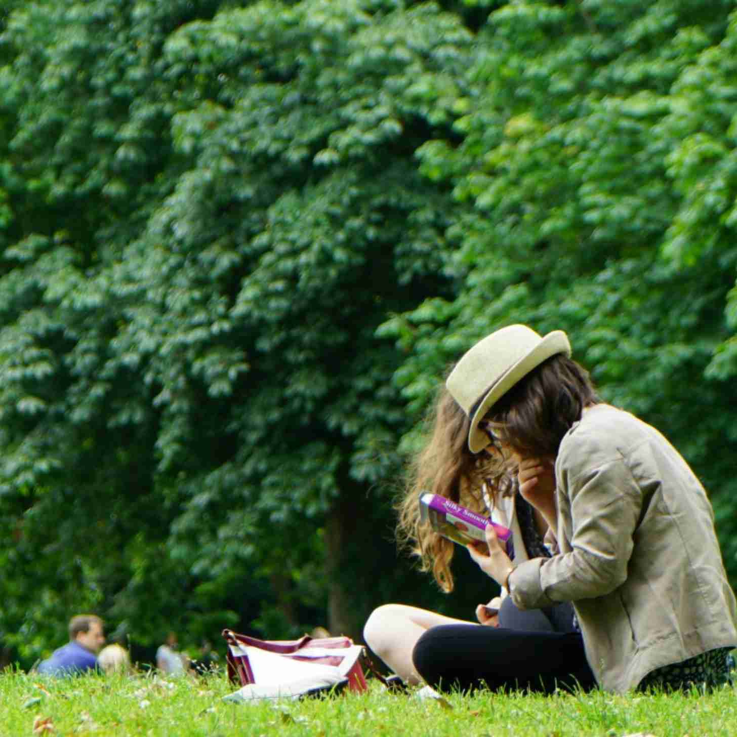 Two women sit in a field in a green, tree-lined park. 