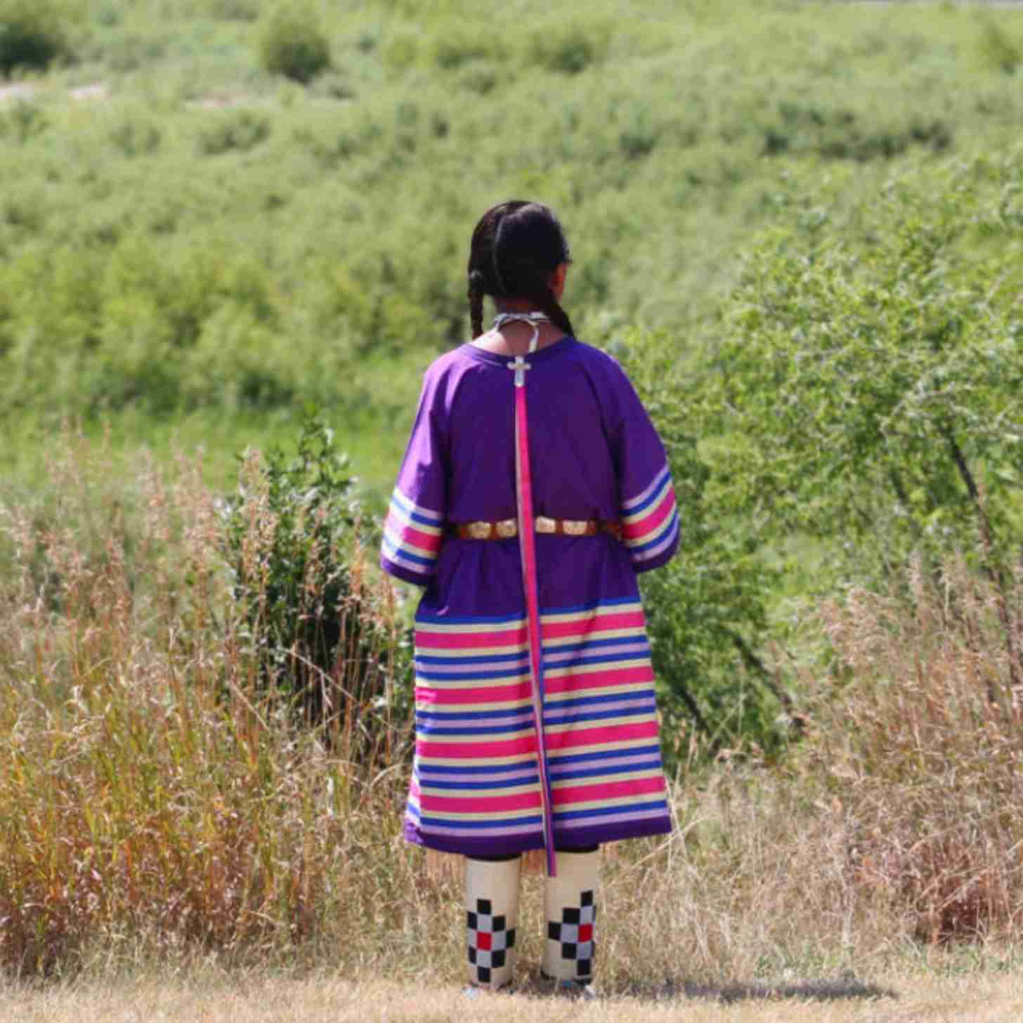A young Native American girl stands at the edge of a green field 