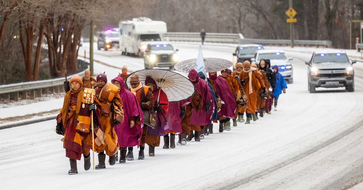 A group of Buddhist monks, dressed in orange robes, walks along the snow-filled streets of North Carolina, escorted by local authorities