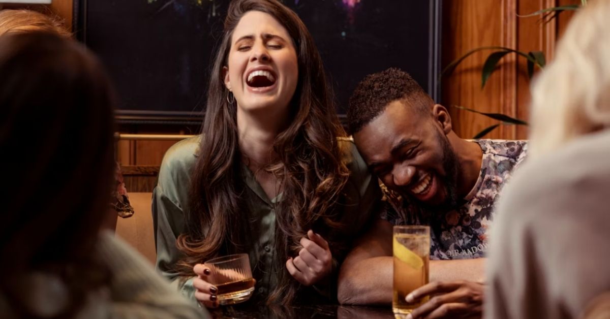 A woman and man laugh at a bar together, drinking whiskey.