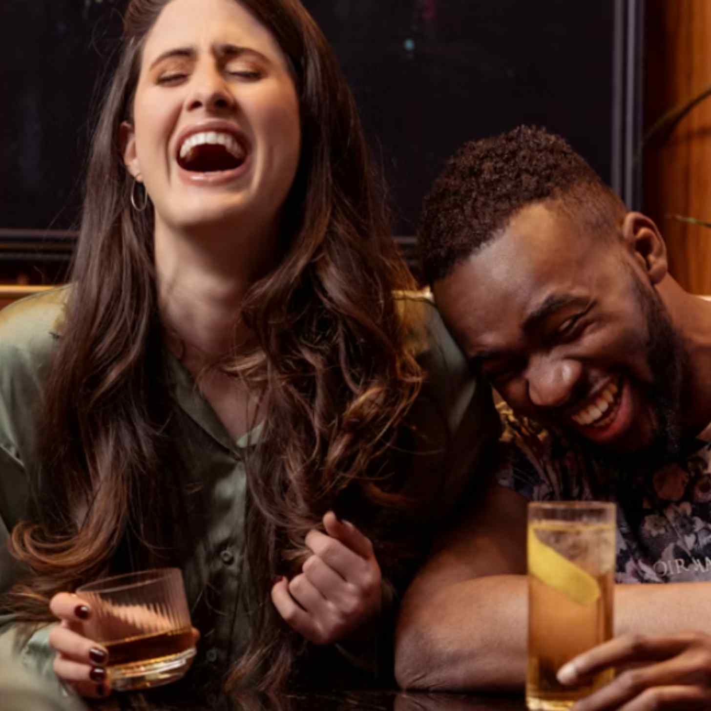 A woman and man laugh at a bar together, drinking whiskey.