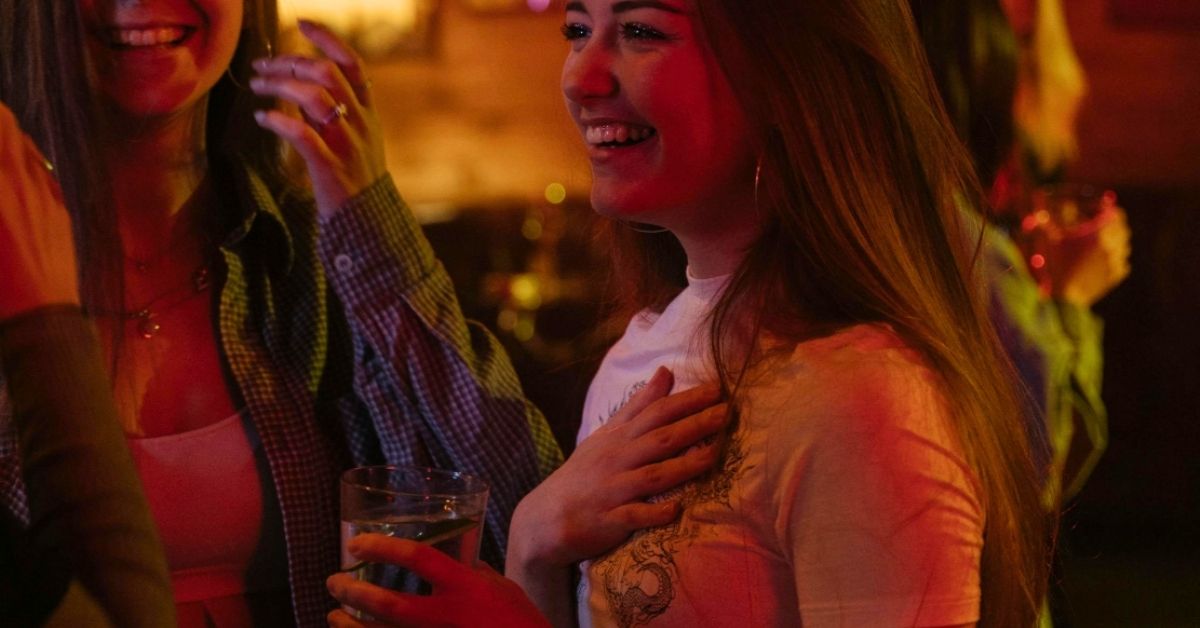 Two women laughing at a bar, as one of them drinks a cup of water.