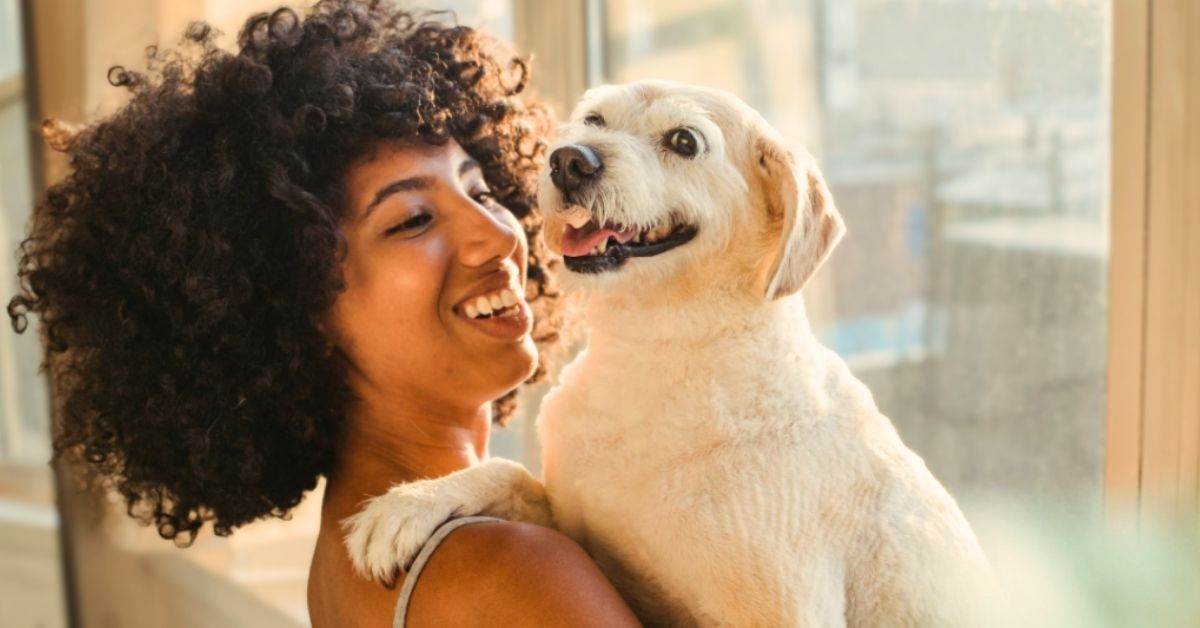 A Black woman smiles while holding a medium sized white dog.