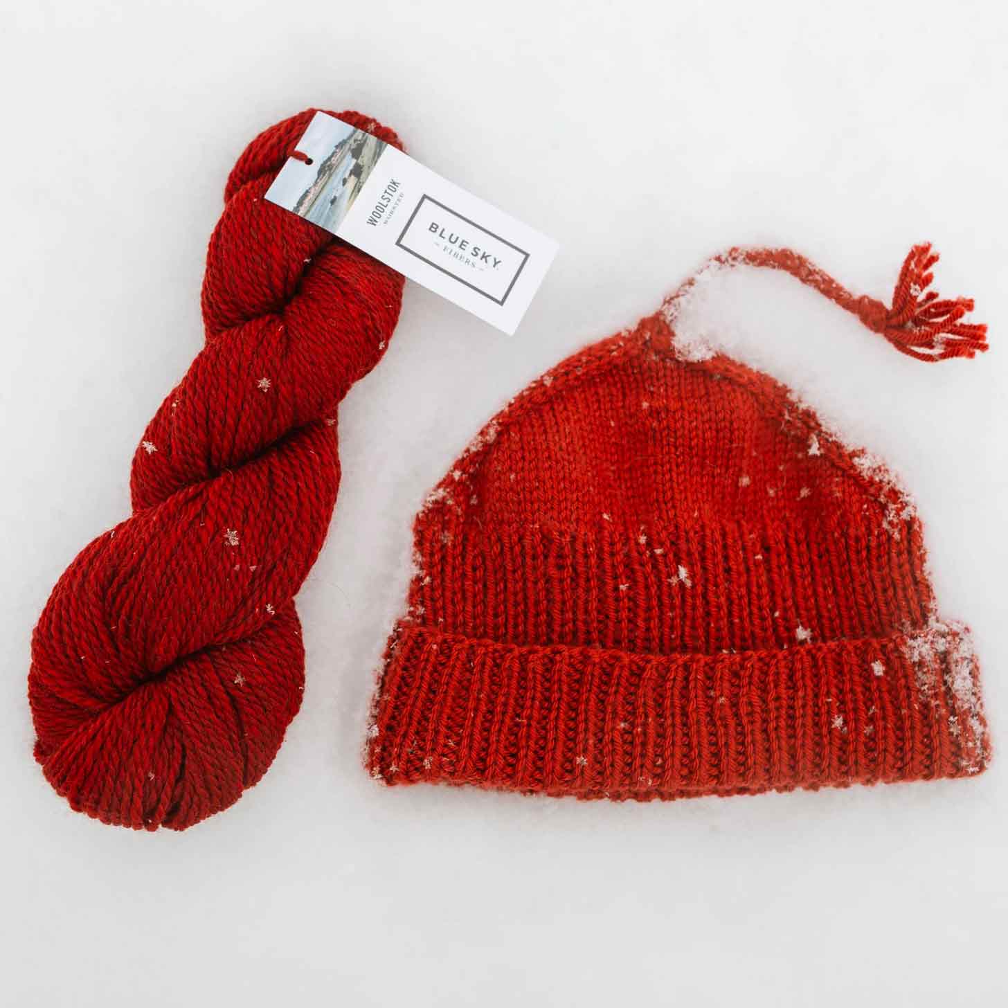 Red "Melt the ICE" knitted hat sits in the snow next to a skein of red yarn