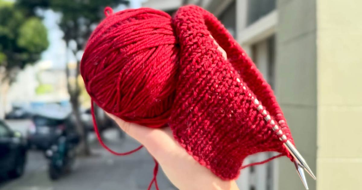 A hand holds up an in-progress red knitted cap and a ball of red yarn
