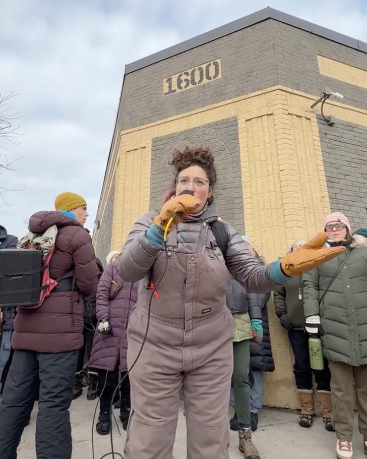A woman in winter gear leads a group of singers outdoors in Minnesota, holding a microphone in mitten-covered hands