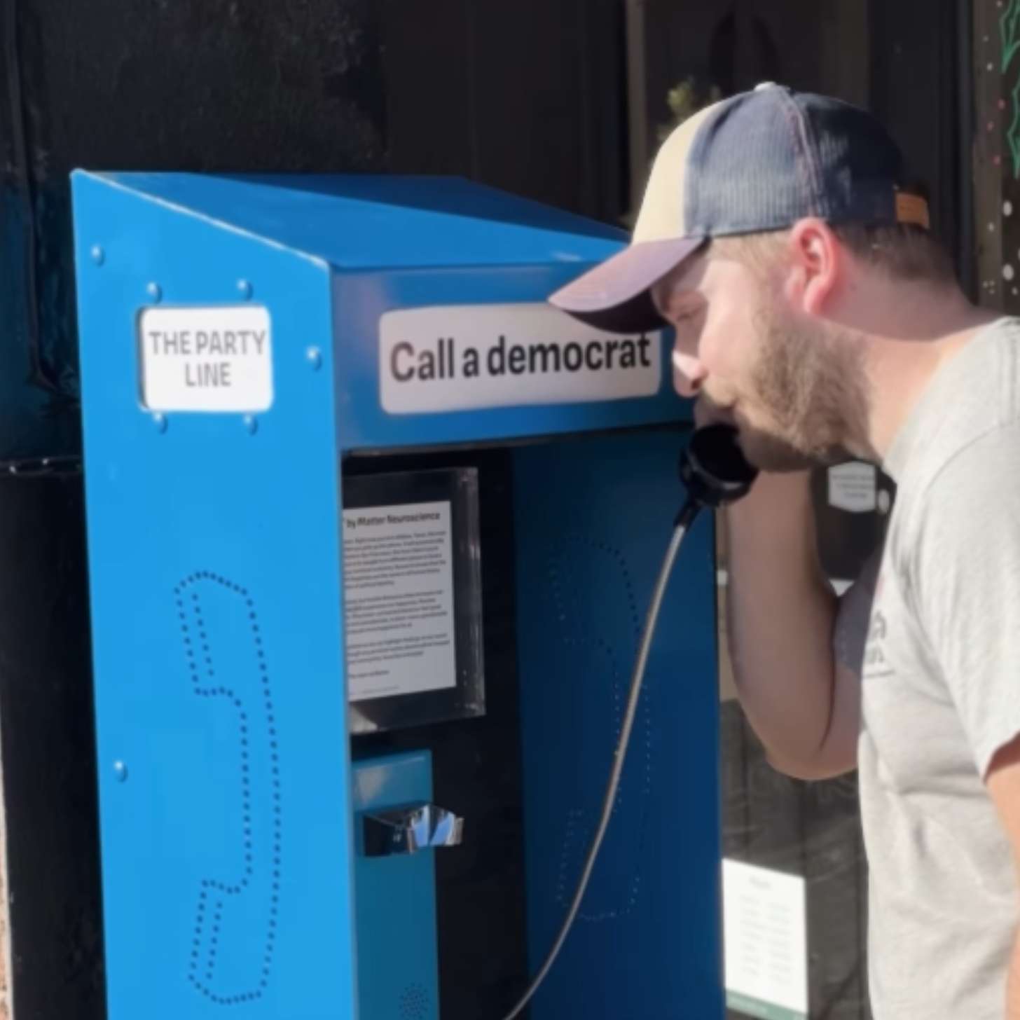 A man with a ball cap talks on a blue payphone on a street corner.