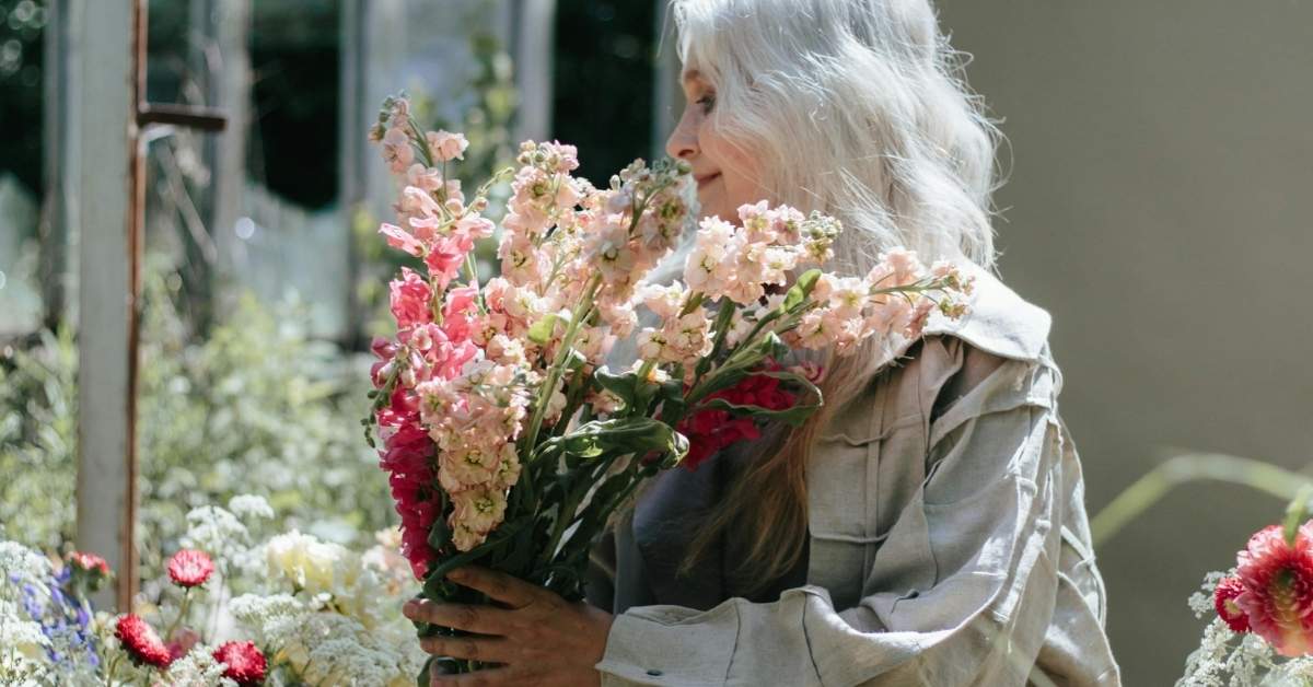 An elderly white woman holds a bouquet of flowers in a greenhouse. 