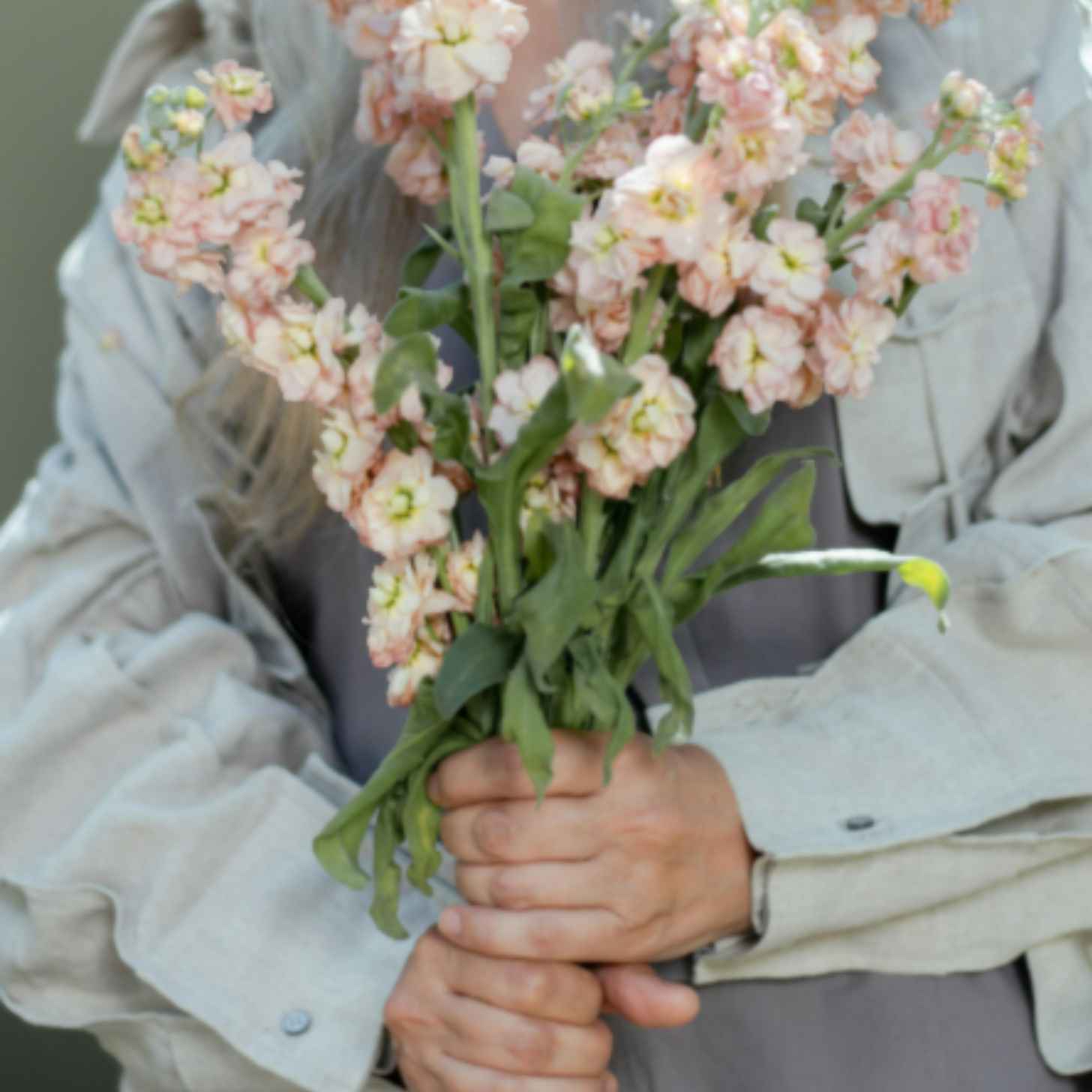 Photographed at chest level, an elderly white woman holds a bouquet of pink flowers. 