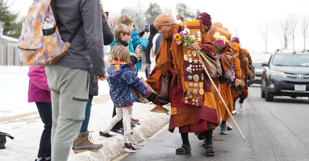 19 Buddhist monks in orange robes walk down a street lined with people. The leader of the group bends down to greet a child while holding flowers in his hands