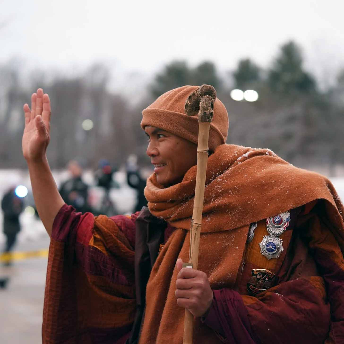 A Buddhist monk in all orange waves to a crowd of people while holding a walking stick, snow falling on his soulders