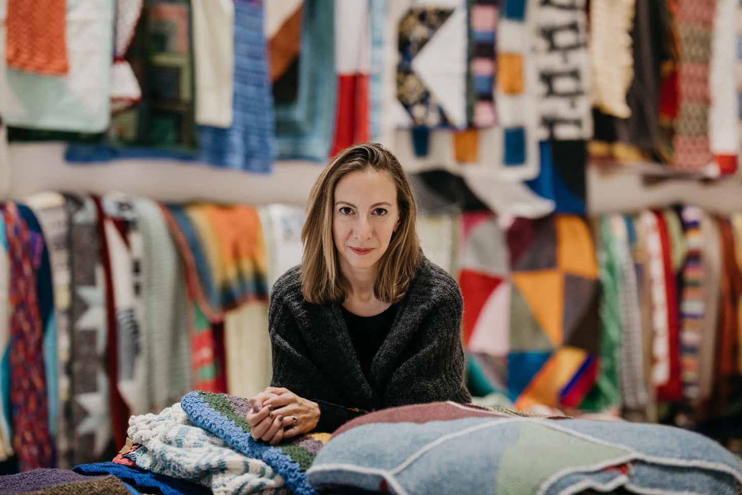A woman with shoulder-length blond hair sits among dozens of colorful handmade blankets