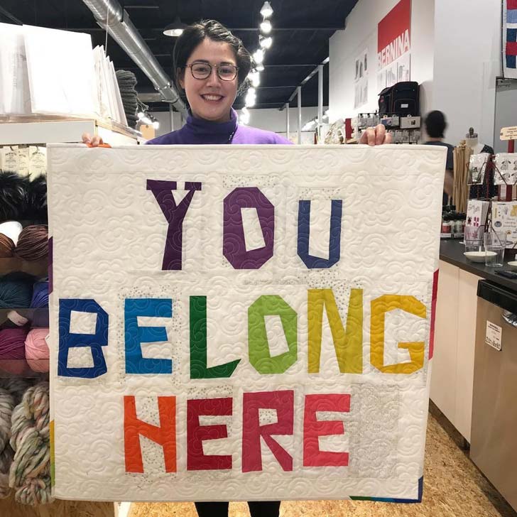 A woman holds up a white quilted blanket with rainbow letters reading "you belong here"