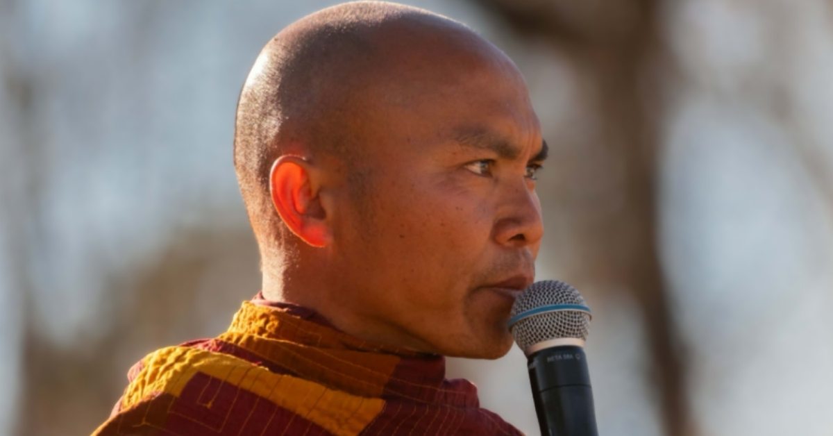 A Buddhist monk in orange robe speaks into a microphone.