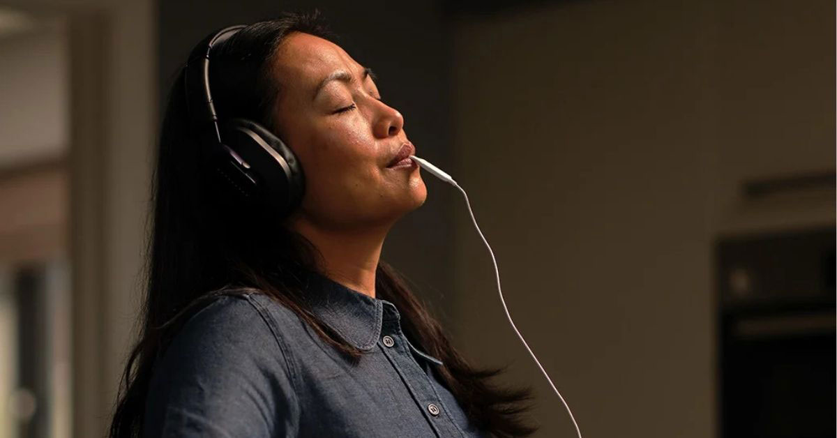 A woman wears headphones over her ears with a white wire coming out of her mouth. She is using an electric mouthpiece from Lenire to treat tinnitus 