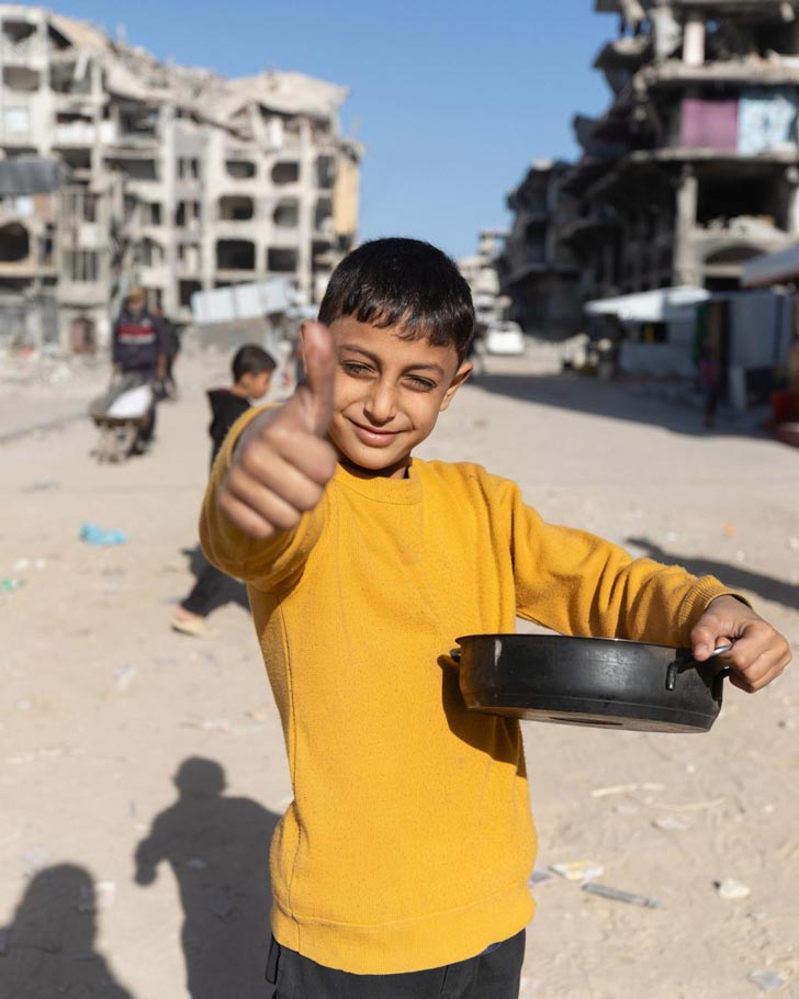 A child in a yellow shirt holds a pot of food in the streets of Gaza. He gives a thumbs-up to the camera.