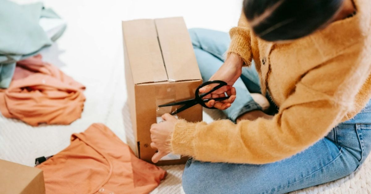 A woman cuts tape off a package as she sits on the floor next to a pile of clothes.