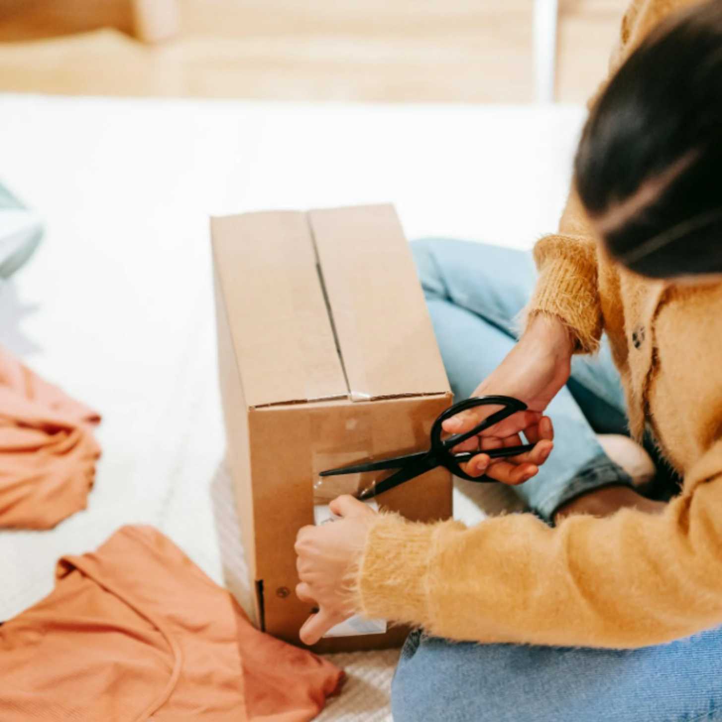 A woman cuts tape off a package as she sits on the floor next to a pile of clothes.