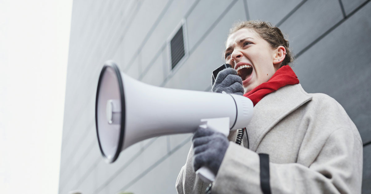 A woman in a white coat, red scarf, and gloves, screams into a megaphone