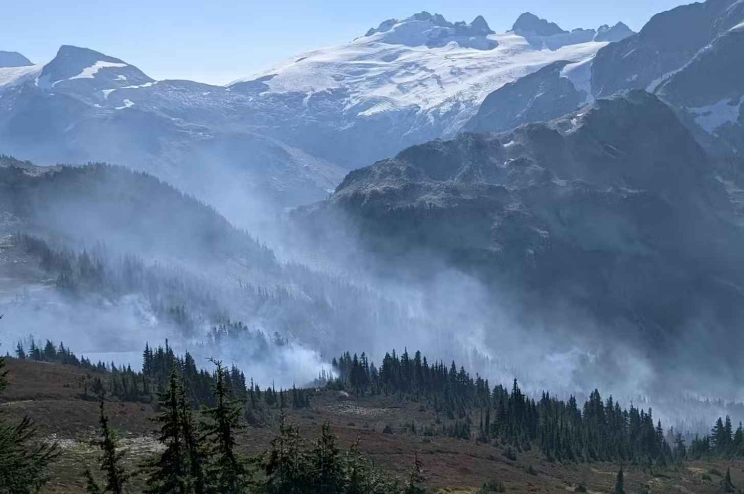 Smoke climbs above a wooded mountainside, with higher peaks in the background.