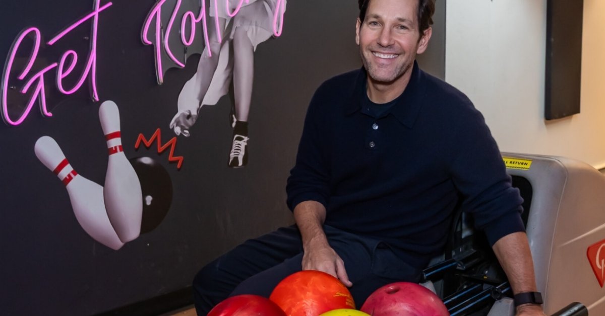 Paul Rudd the actor sits down next to a rack of bowling balls in a bowling alley.
