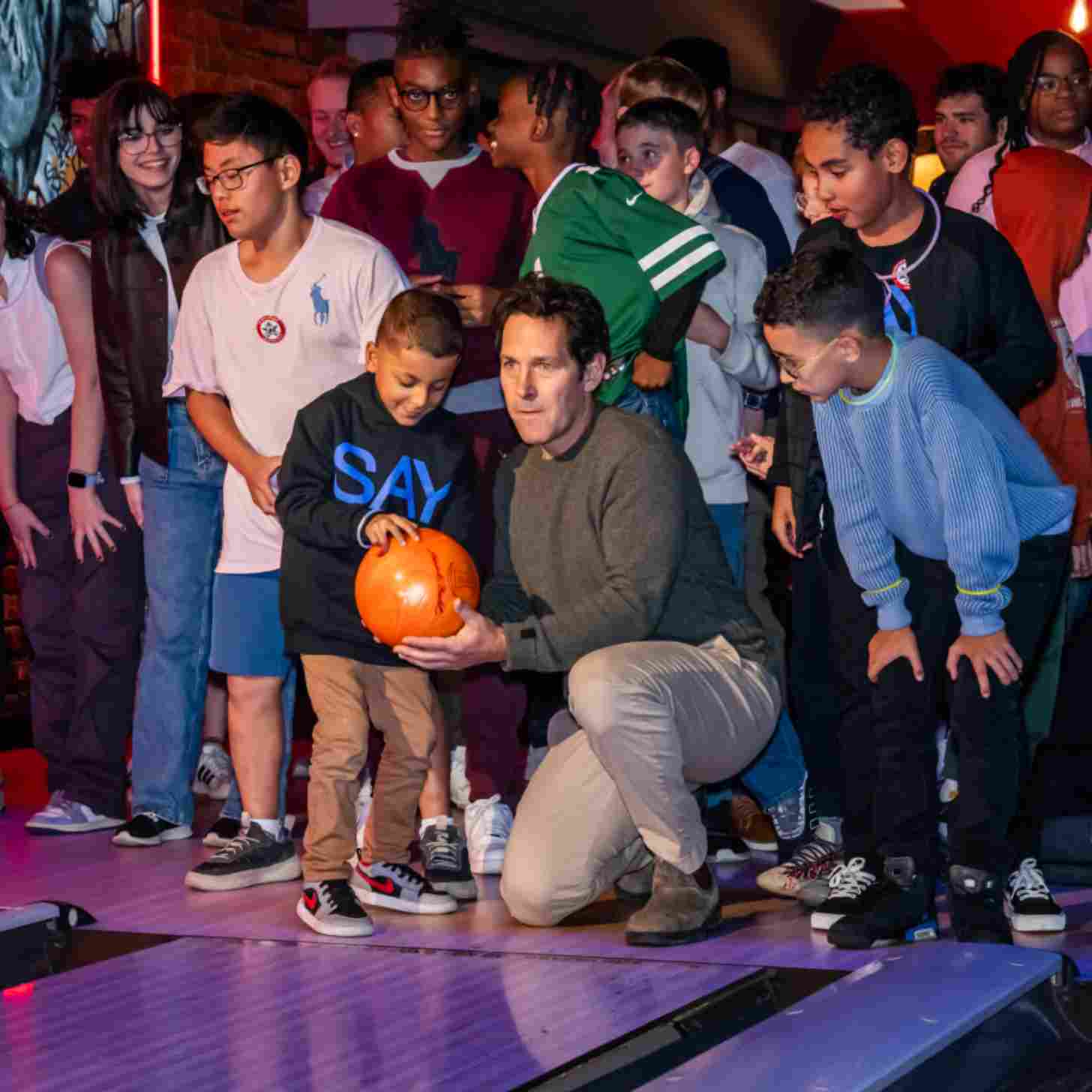 Paul Rudd with a group of young kids and teens in a bowling alley, ready to roll a ball down a lane.